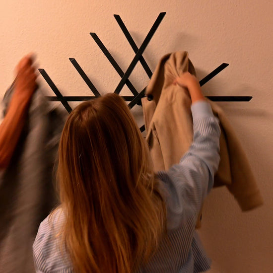 Drei Besucher betreten den Flur und hängen ihre Jacken an die Treecado Wandgarderobe. Das moderne, anthrazitfarbene Designobjekt ist an einer hellen Wand montiert und zeigt, wie einfach Kleidung aufgehängt werden kann.